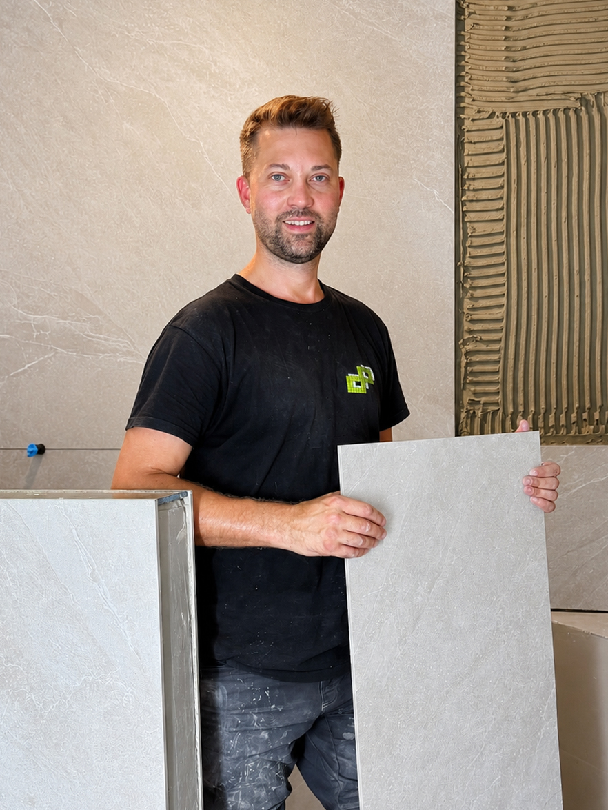 Daniel Pätzug lächelt und hält eine großformatige Fliese vor einer Wand mit frisch aufgetragenem Fliesenkleber