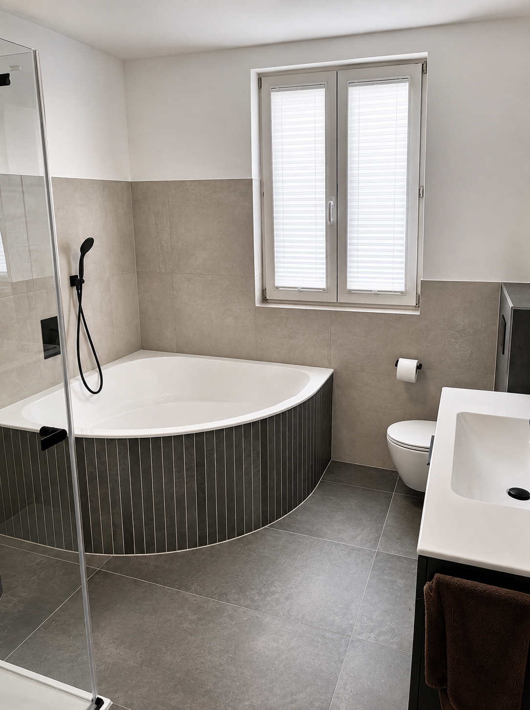 Eckbadewanne mit abgerundeter, dunkel gefliester Verkleidung neben Fenster, WC und Waschtisch
