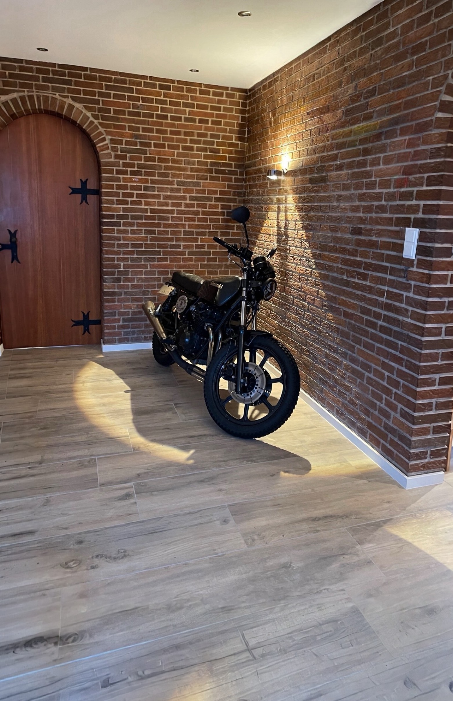 Raumecke mit Backsteinwand, gewölbter Holztür und Bodenfliesen in Holzoptik, davor ein geparktes Motorrad