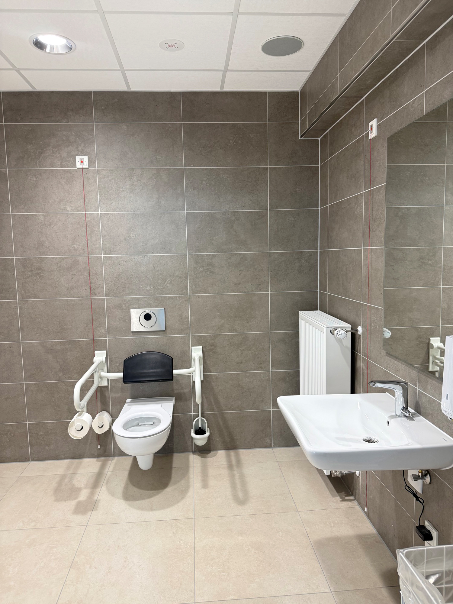 Barrierefreies WC mit grauen Wandfliesen, Klappgriffen und wandhängendem Waschbecken